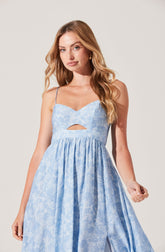 A woman is wearing a light blue sleeveless dress with thin spaghetti straps. The dress has a sweetheart neckline with a small keyhole cutout just below the bust. The fabric features a subtle pattern, and the skirt is gathered, creating a flowy, gathered silhouette.